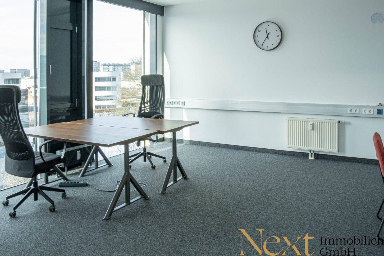 Funktionales Büro mit großem Fenster, Heizkörper und Uhr an der Wand, bietet viel Tageslicht.
