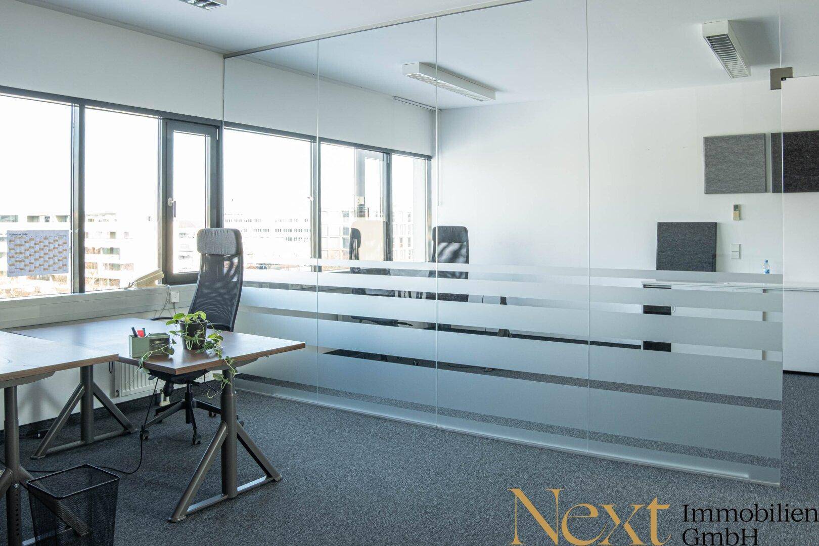 Modernes Büro mit Glaswand als Raumteiler, großen Fenstern und Teppichboden, schafft offene Atmosphäre.
