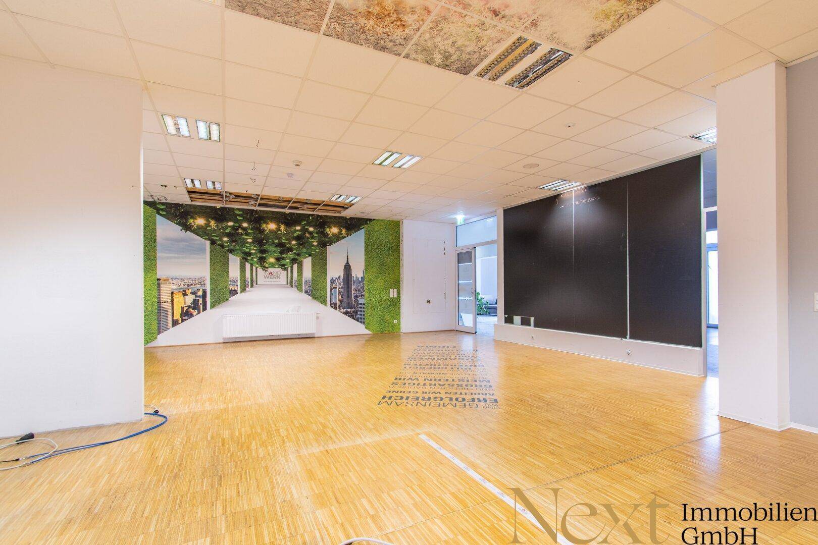 Weitläufiger Bürobereich mit Holzparkettboden, dekorativen Wandbildern und einer abgehängten Decke, die vielseitige Nutzungsmöglichkeiten bietet.