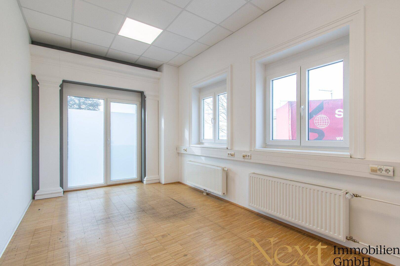 Helles Büro mit Holzparkettboden, großen Fenstern und Heizkörpern, das eine angenehme Arbeitsatmosphäre bietet.