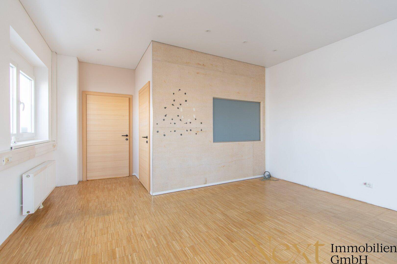Schlichter Büroraum mit Holzparkettboden, einer Holzwand und zwei Türen, der vielseitig nutzbar ist.