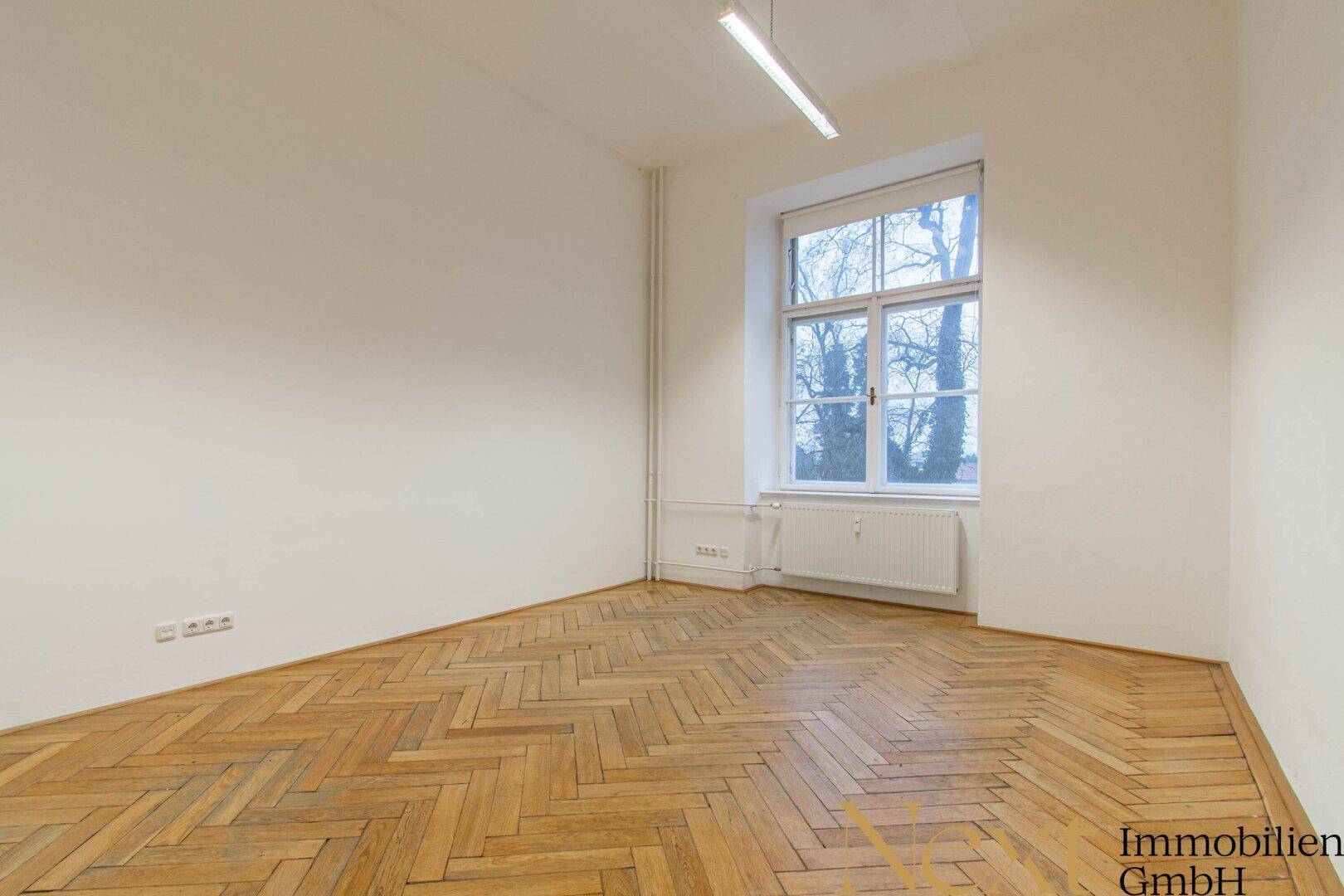Heller, leerer Raum mit Fischgrätparkett, einem großen Fenster und einem Heizkörper unter der Fensterbank.