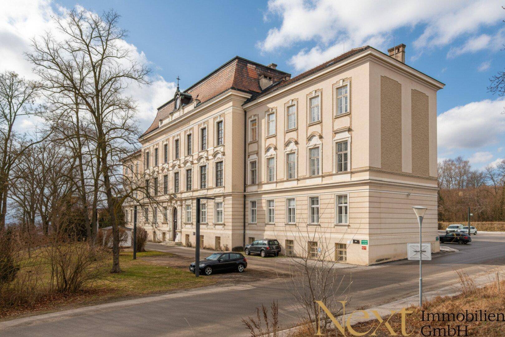 Historisches Gebäude mit klassischer Architektur, umgeben von Bäumen und einem Parkplatz unter blauem Himmel.