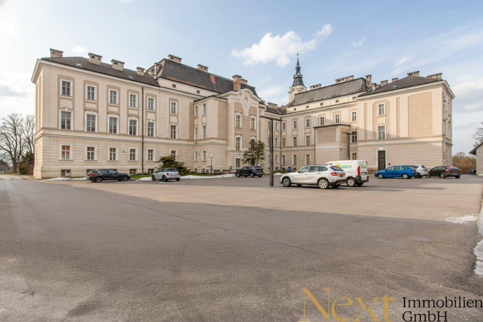 Großes historisches Gebäude mit weitläufigem Parkplatz und klassischer Architektur unter einem sonnigen Himmel.