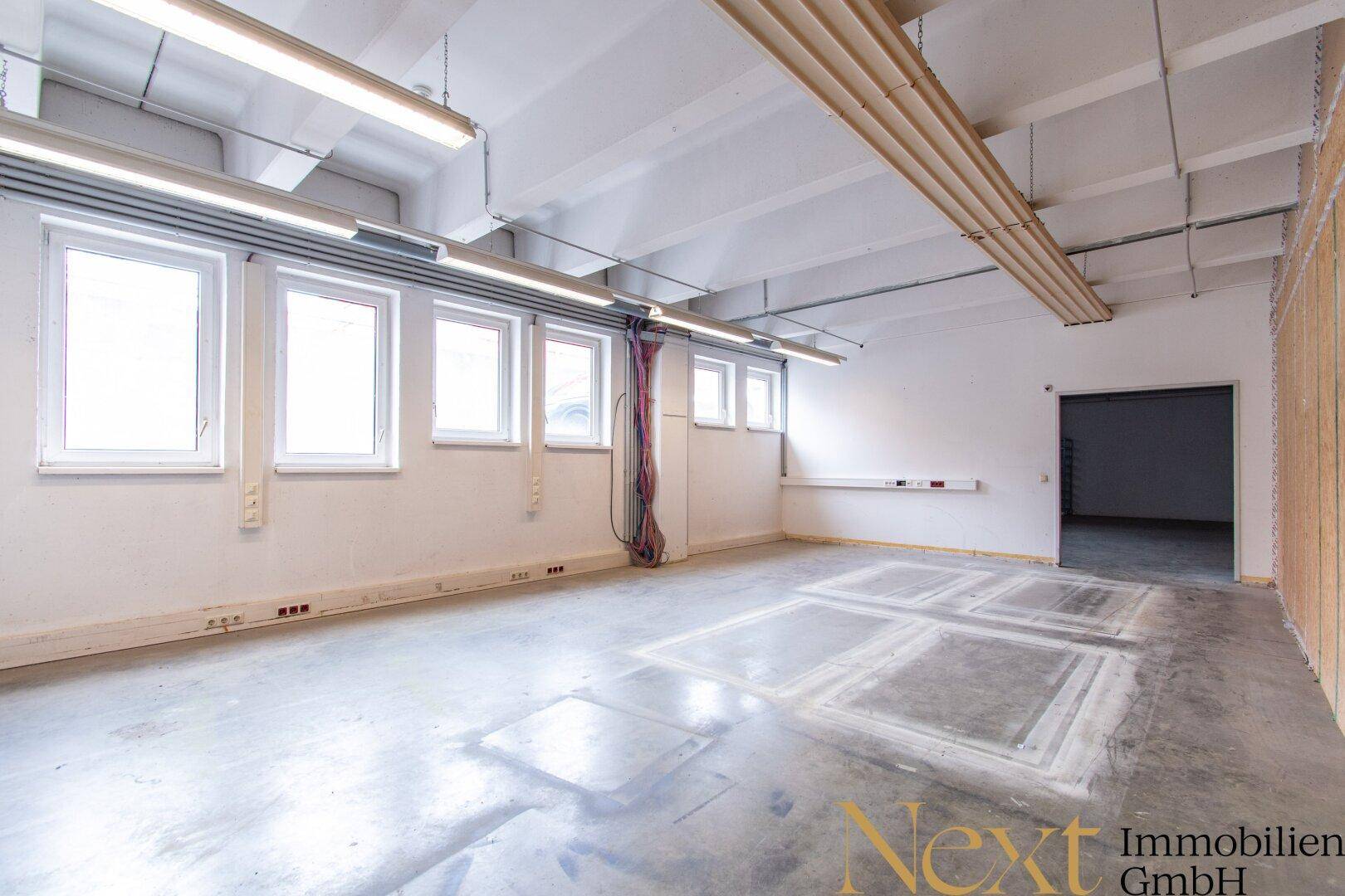 Heller Gewerberaum mit großen Fenstern, Betonboden und sichtbaren Deckeninstallationen, bietet viel Tageslicht.