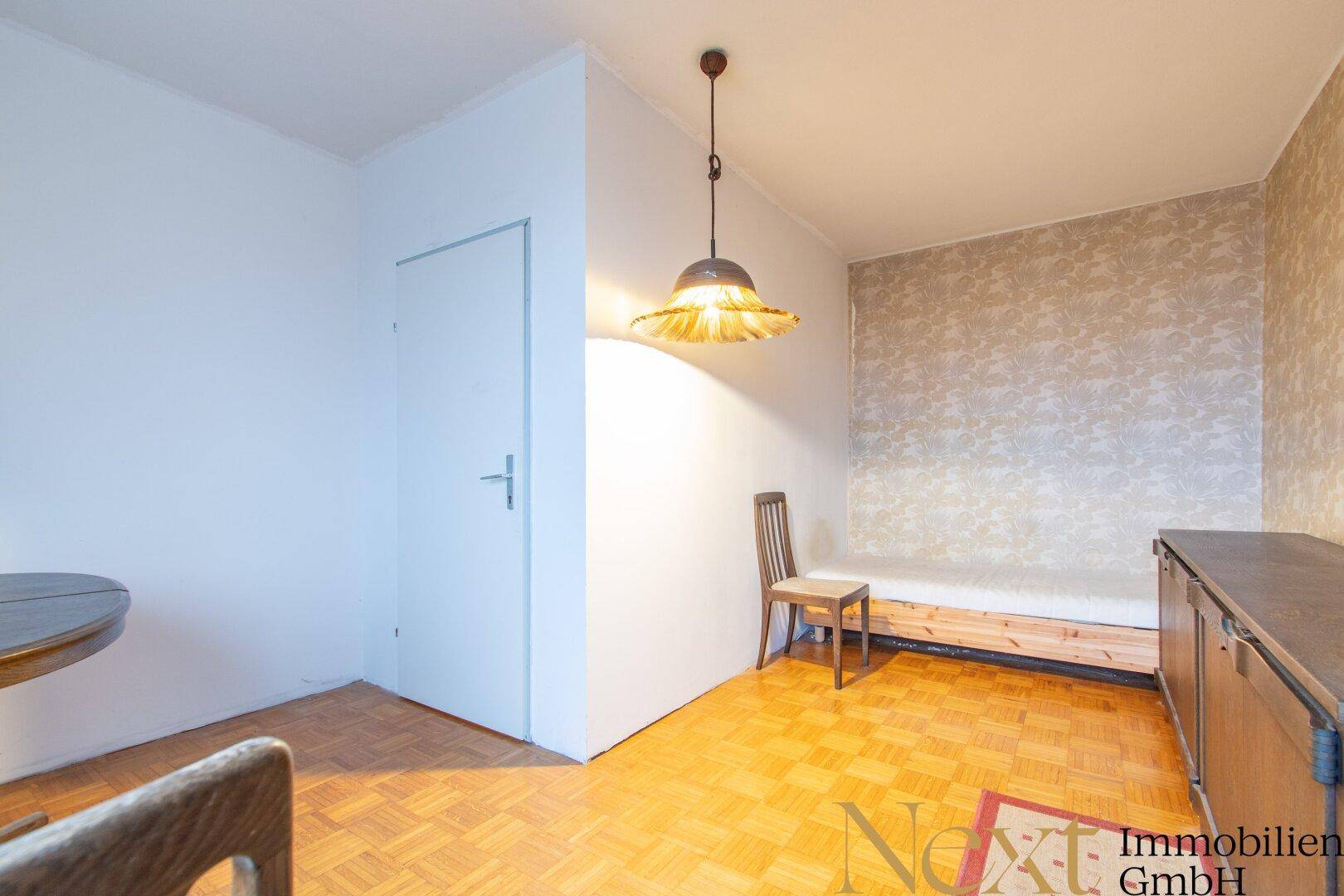 Helles Zimmer mit Parkettboden, einfacher Einrichtung und einer gemusterten Akzentwand.
