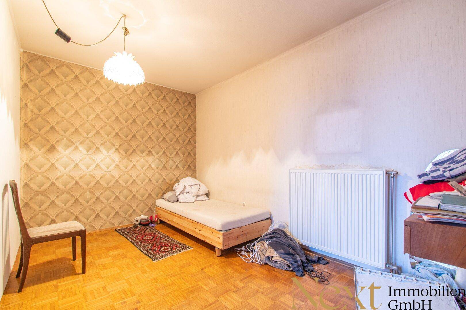 Schlafzimmer mit Parkettboden, Heizkörper und einer gemusterten Tapetenwand.