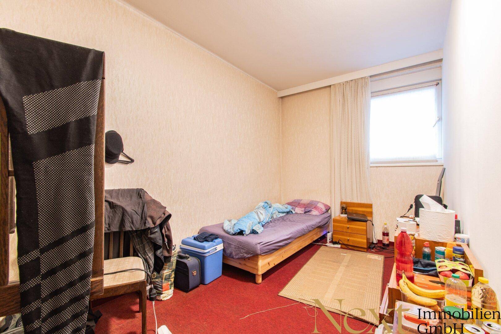 Schlafzimmer mit rotem Teppichboden, Fenster und einfacher Einrichtung.