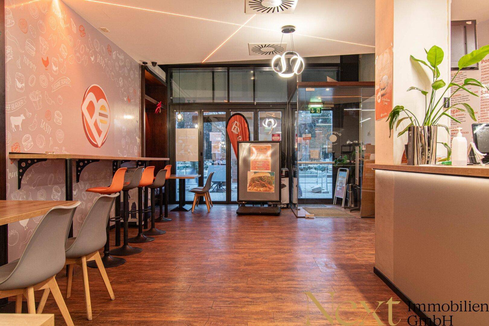 Eingangsbereich eines Restaurants mit großen Glasfronten, Thekenbestuhlung und modernem Interieur.