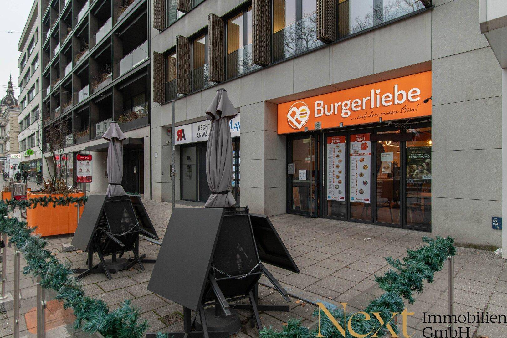 Moderne Geschäftsfassade mit großen Fenstern, Restaurant-Eingang und Außenbestuhlung auf dem Bürgersteig.