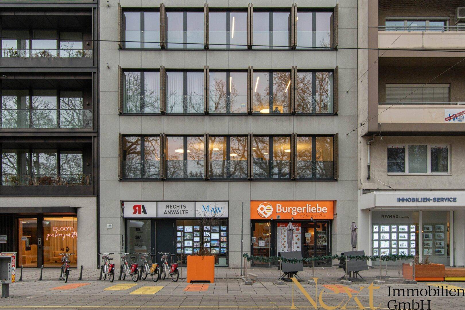 Ansicht eines modernen Geschäftsgebäudes mit mehreren Etagen, großen Fenstern und verschiedenen Ladenfronten.