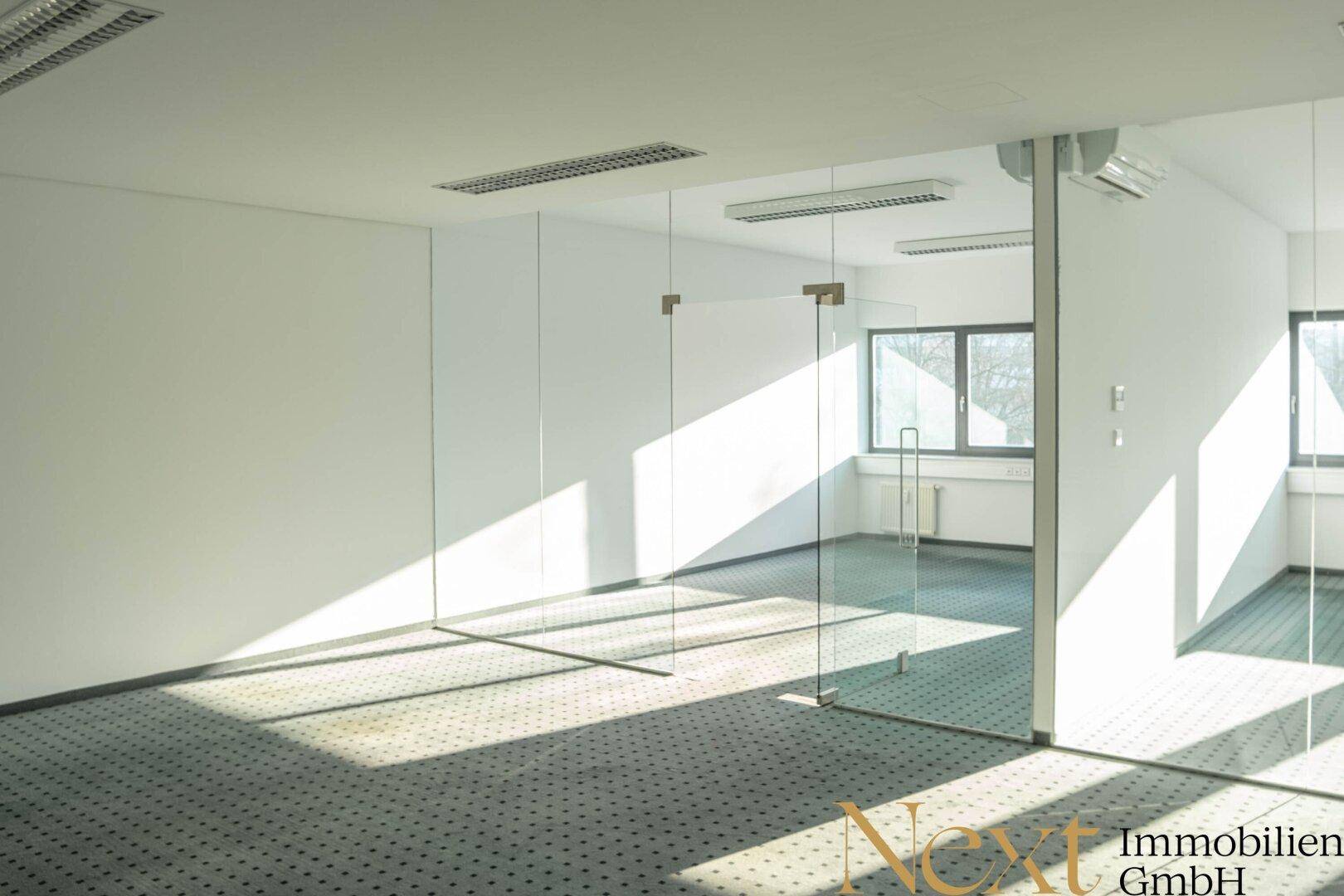 Geräumiges Büro mit Teppichboden, Glasabtrennungen und Fenstern, die viel Sonnenlicht hereinlassen.