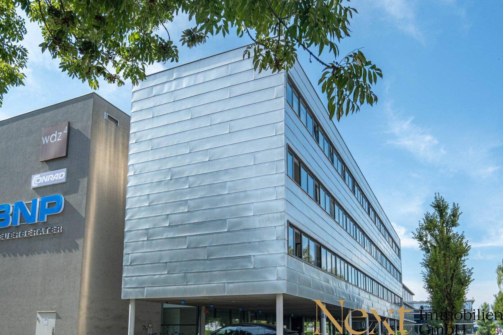 Bürogebäude mit moderner Metallfassade und großen Fenstern, umgeben von Bäumen an einem sonnigen Tag.