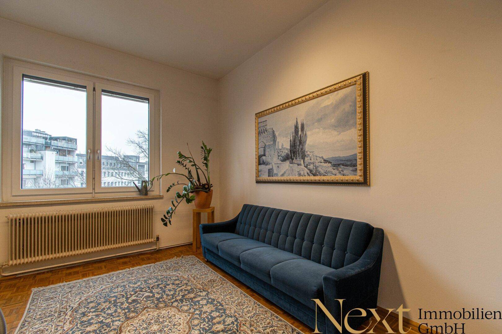 Gemütliches Wohnzimmer mit blauem Sofa, Teppich und Blick auf die Stadt durch das Fenster.