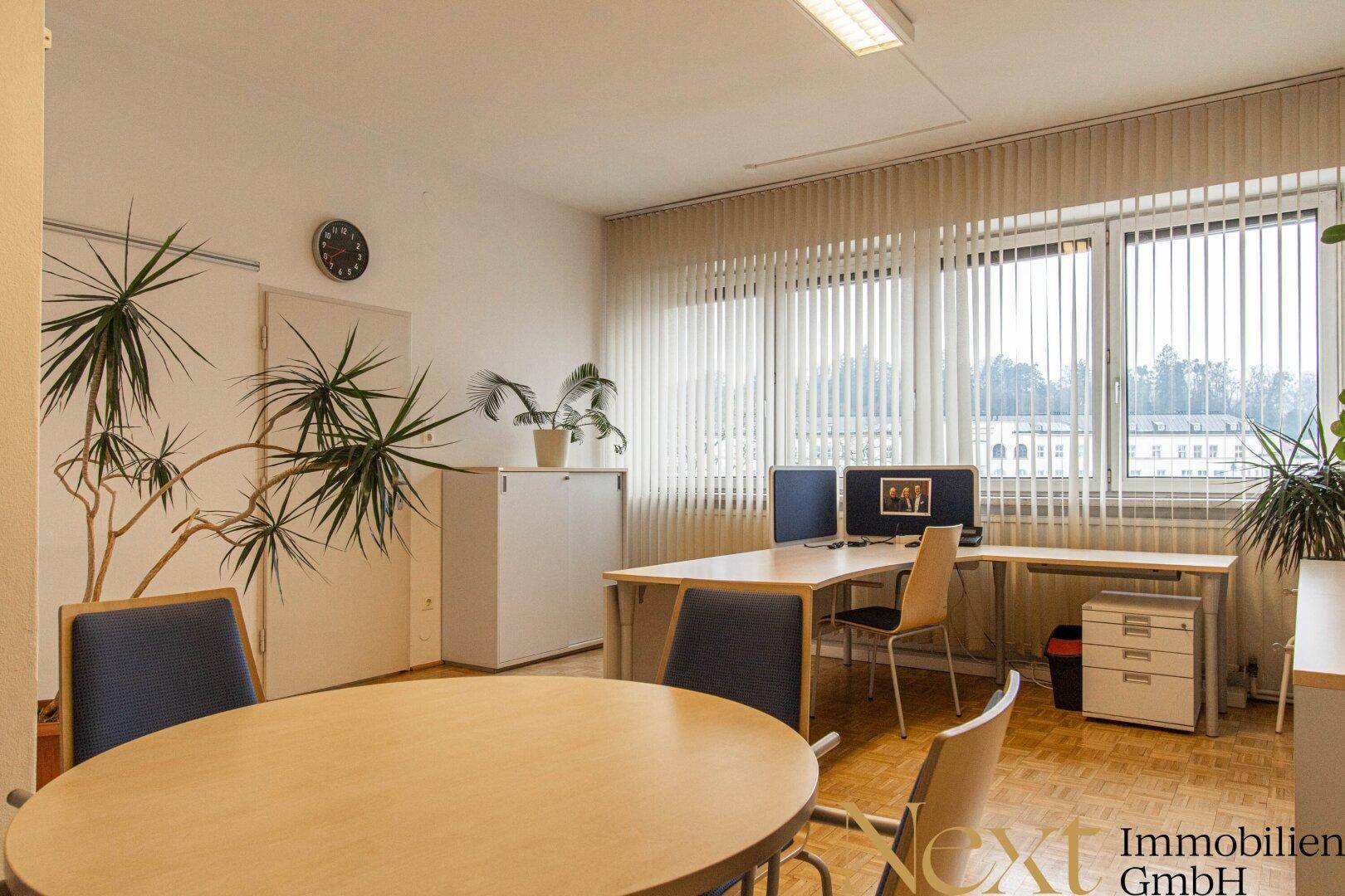 Helles Büro mit mehreren Arbeitsplätzen, Besprechungstisch und großen Fenstern mit Jalousien.