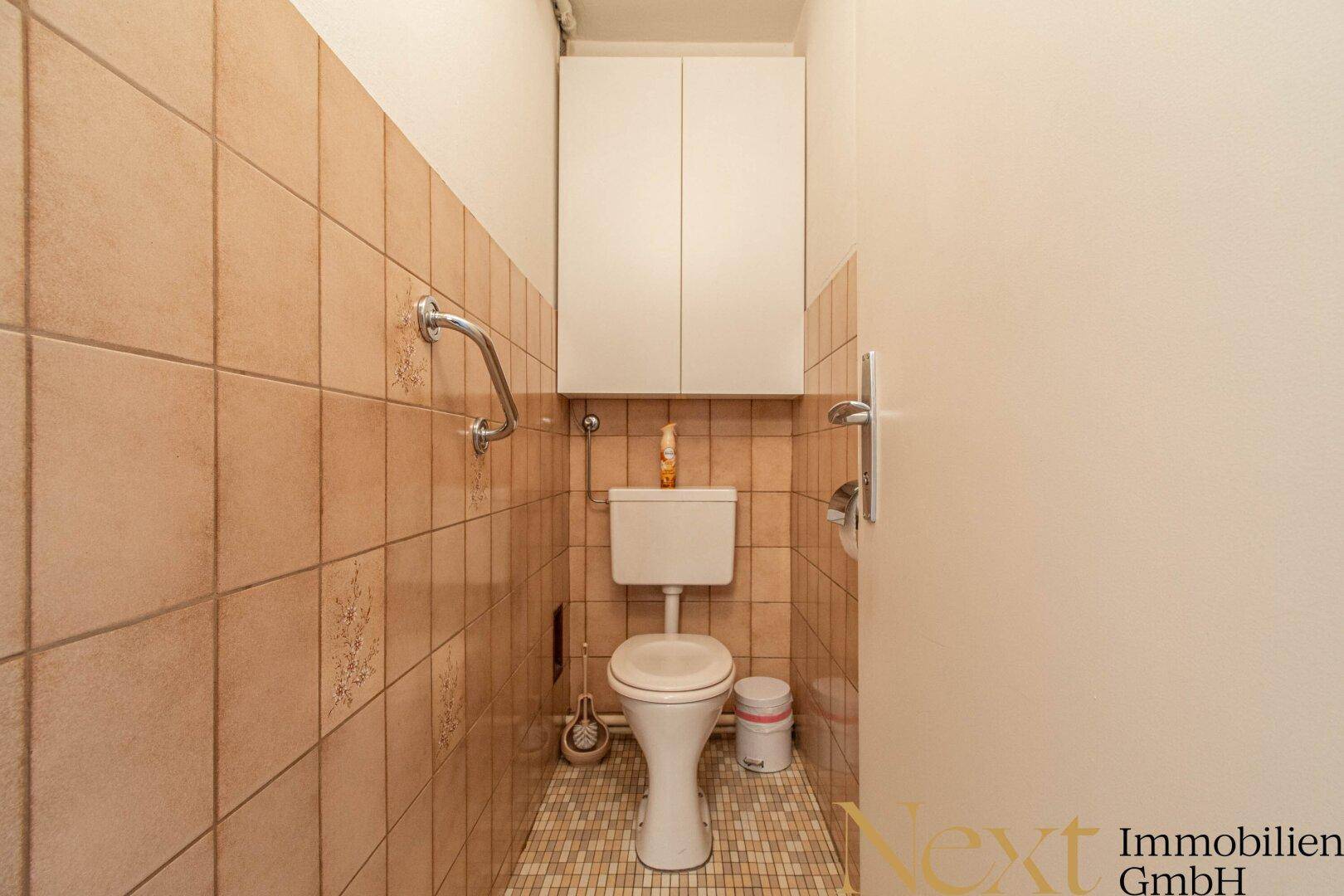 Separate Toilette mit beigefarbenen Fliesen, Haltegriffen und einem kleinen Wandschrank.