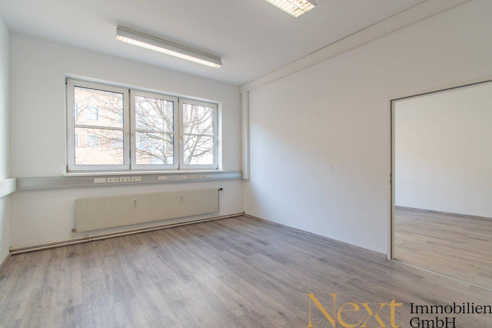Helles Büro mit großen Fenstern, modernem Laminatboden und Zugang zu einem weiteren Raum.