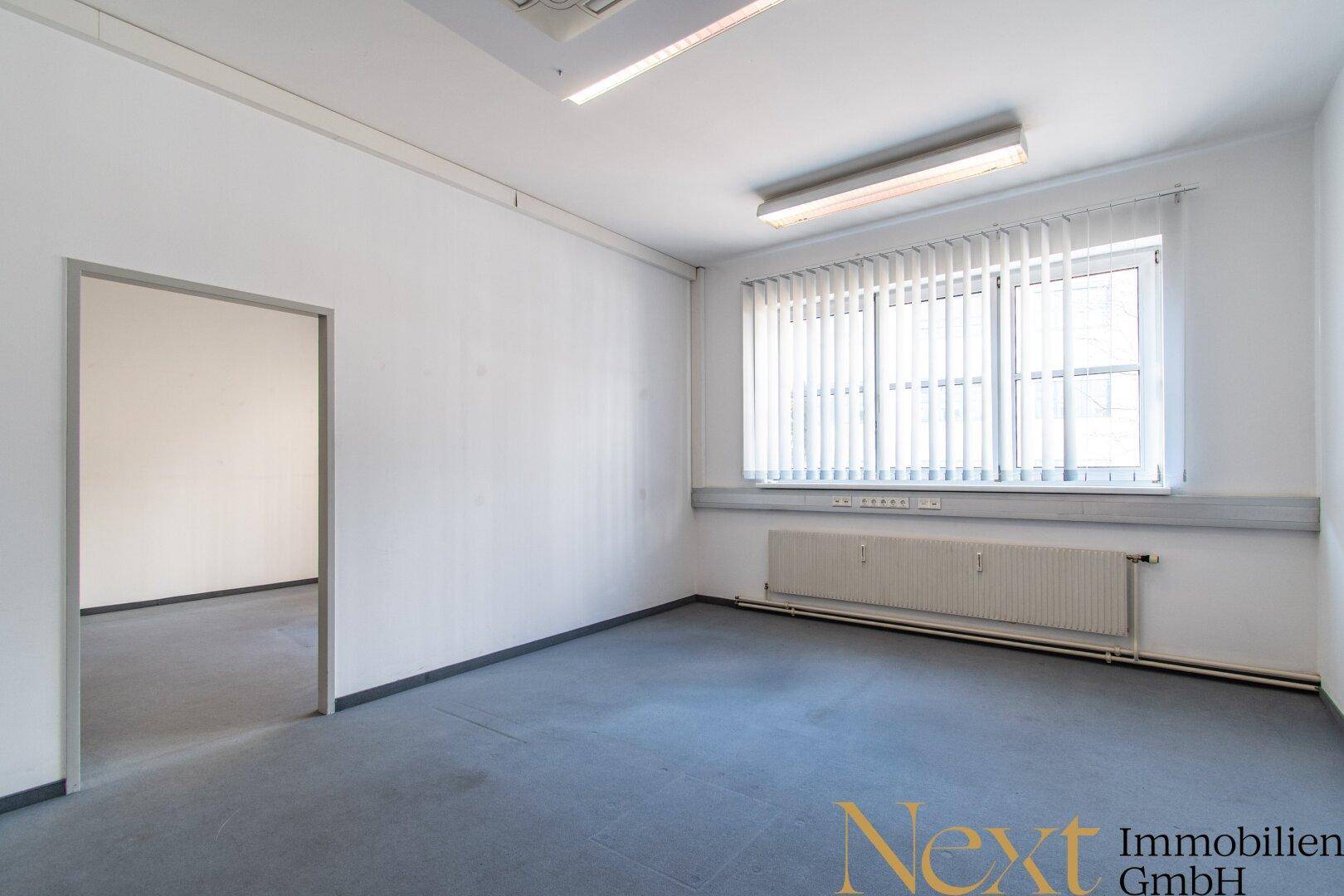 Bürofläche mit Teppichboden, Fenster mit Jalousien und Zugang zu einem weiteren Raum.