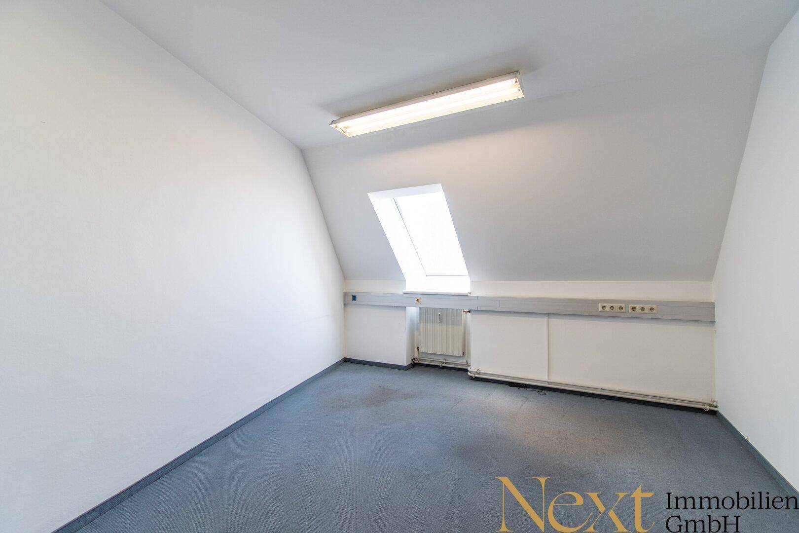 Heller Büroraum im Dachgeschoss mit einem Dachfenster, schrägen Wänden und Teppichboden.