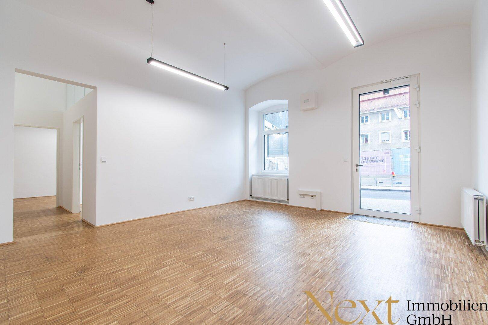 Heller, leerer Raum mit Holzboden, großem Fenster und einer Glastür zum Außenbereich.