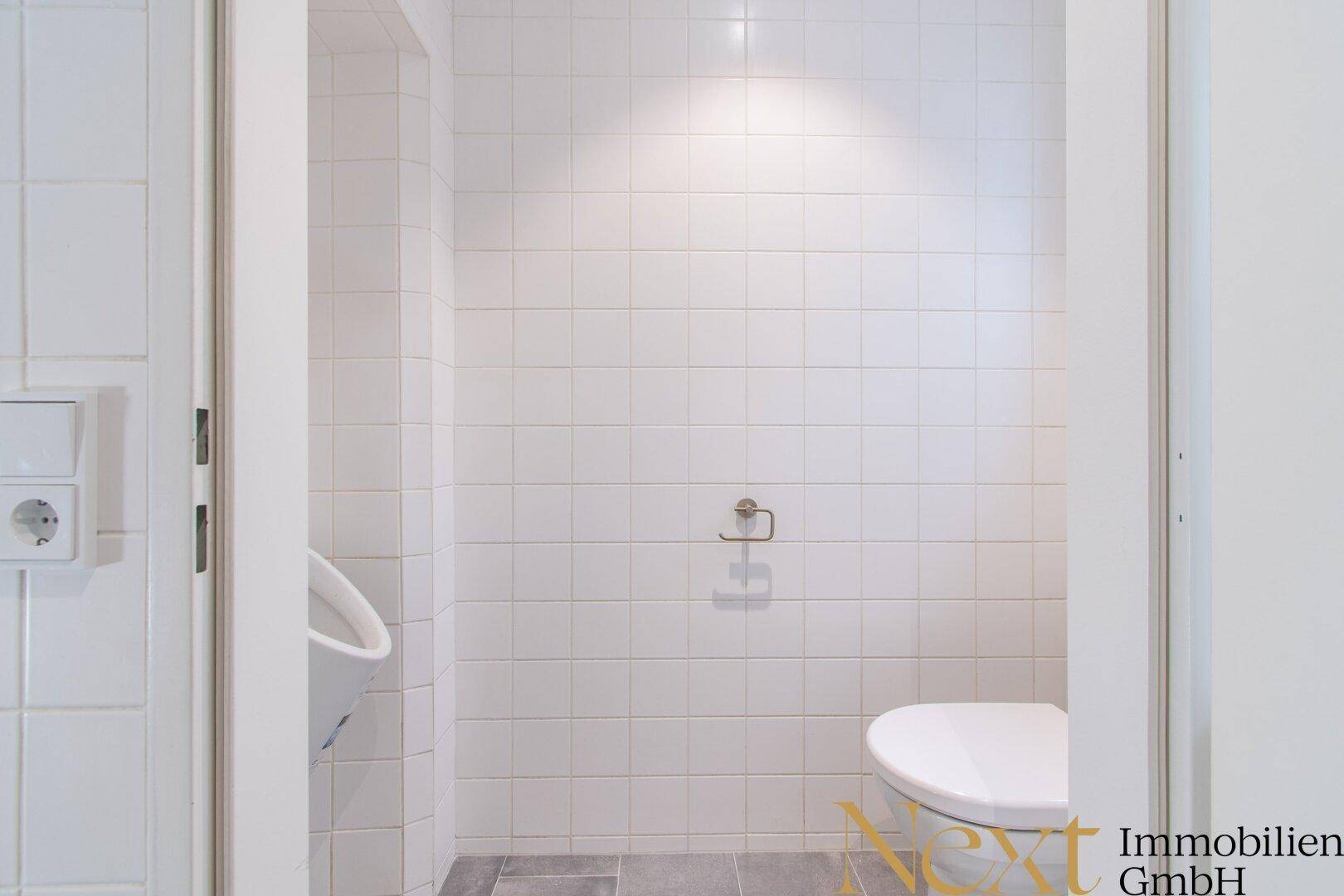 Separate Toilette mit Urinal und WC, komplett gefliest in Weiß mit dunklem Boden.