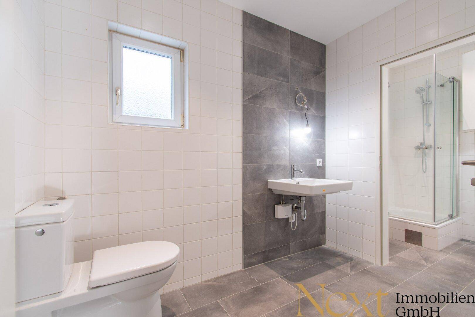 Geräumiges Badezimmer mit WC, Waschbecken und einer Duschkabine, hell gefliest mit dunklen Akzenten.