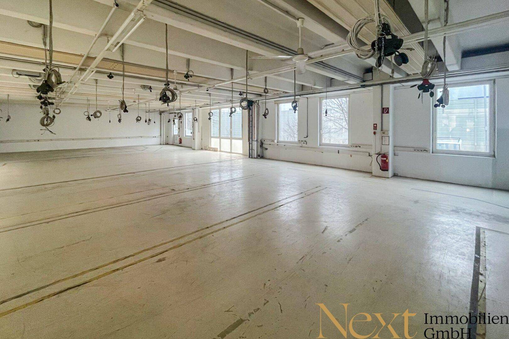 Geräumiger, heller Gewerberaum mit großen Fenstern, Betonboden und zahlreichen Deckeninstallationen für flexible Nutzung.