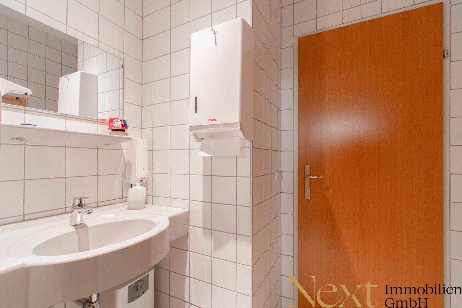 Modernes Badezimmer mit Waschbecken, Spiegel und weißen Fliesen, ausgestattet mit einem Handtuchspender.
