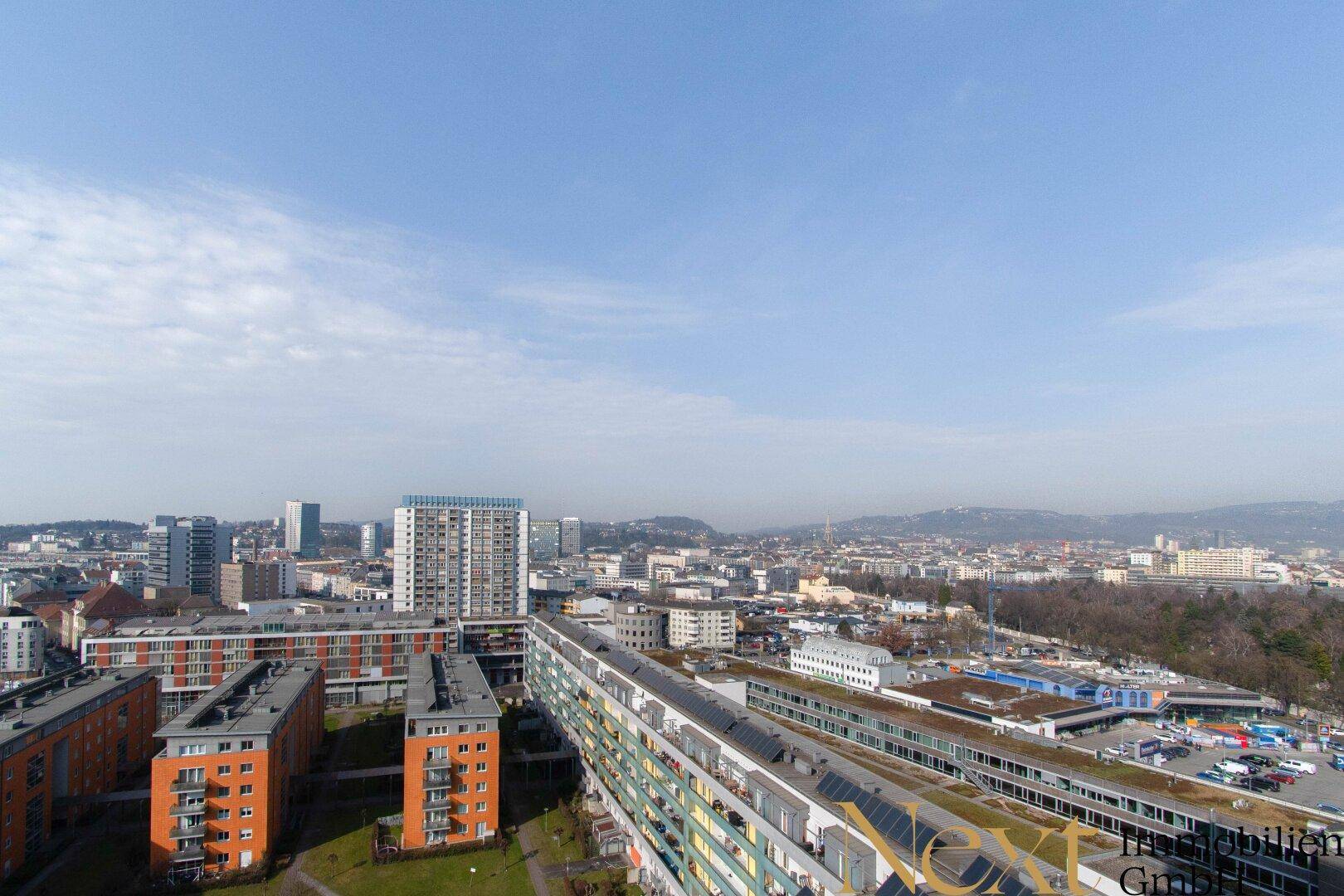 Weitläufige Luftaufnahme einer Stadtlandschaft mit Wohngebäuden, Grünflächen und Bergen im Hintergrund.