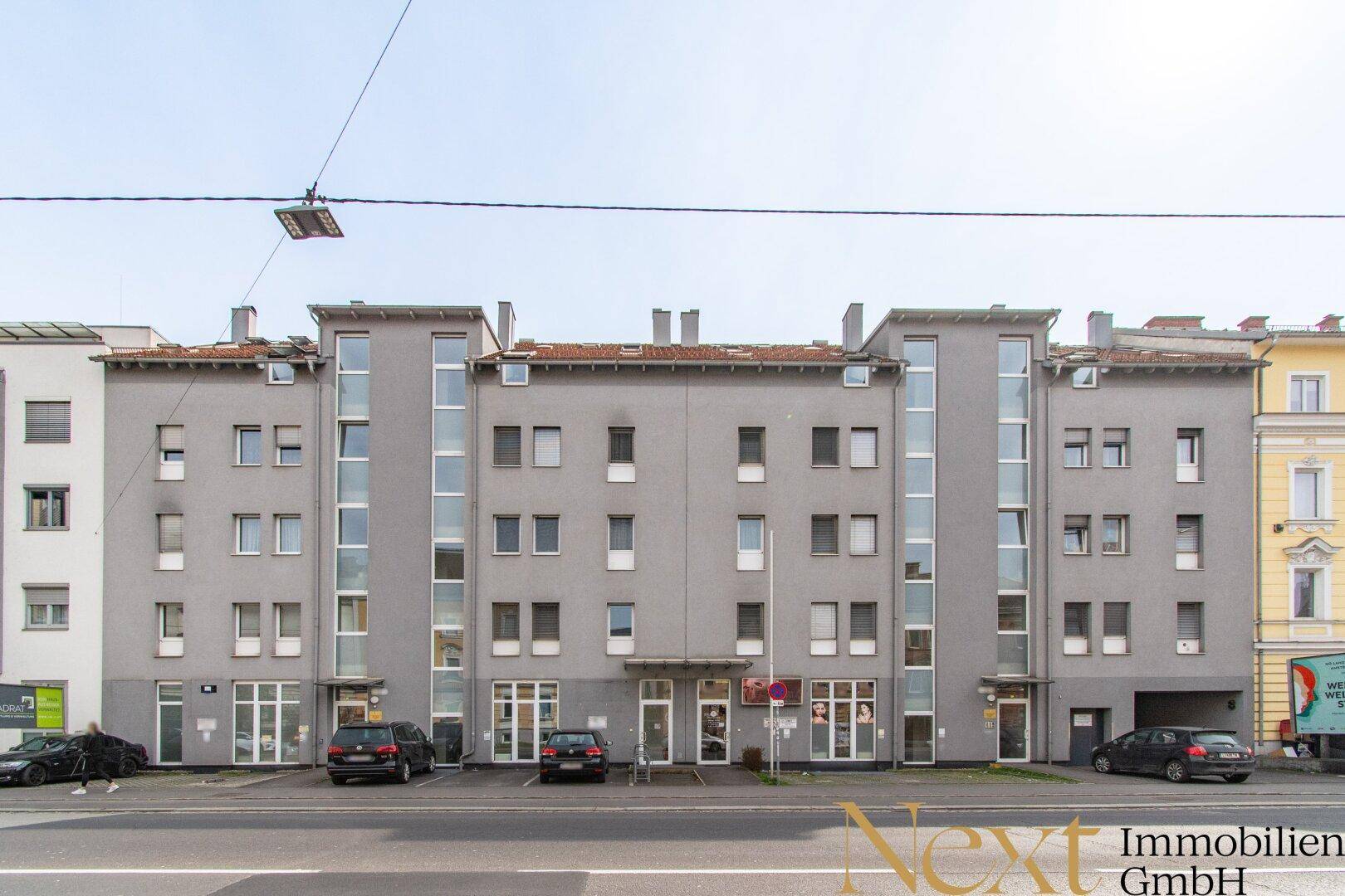 Mehrstöckiges Gebäude mit grauer Fassade, vielen Fenstern und Geschäften im Erdgeschoss.
