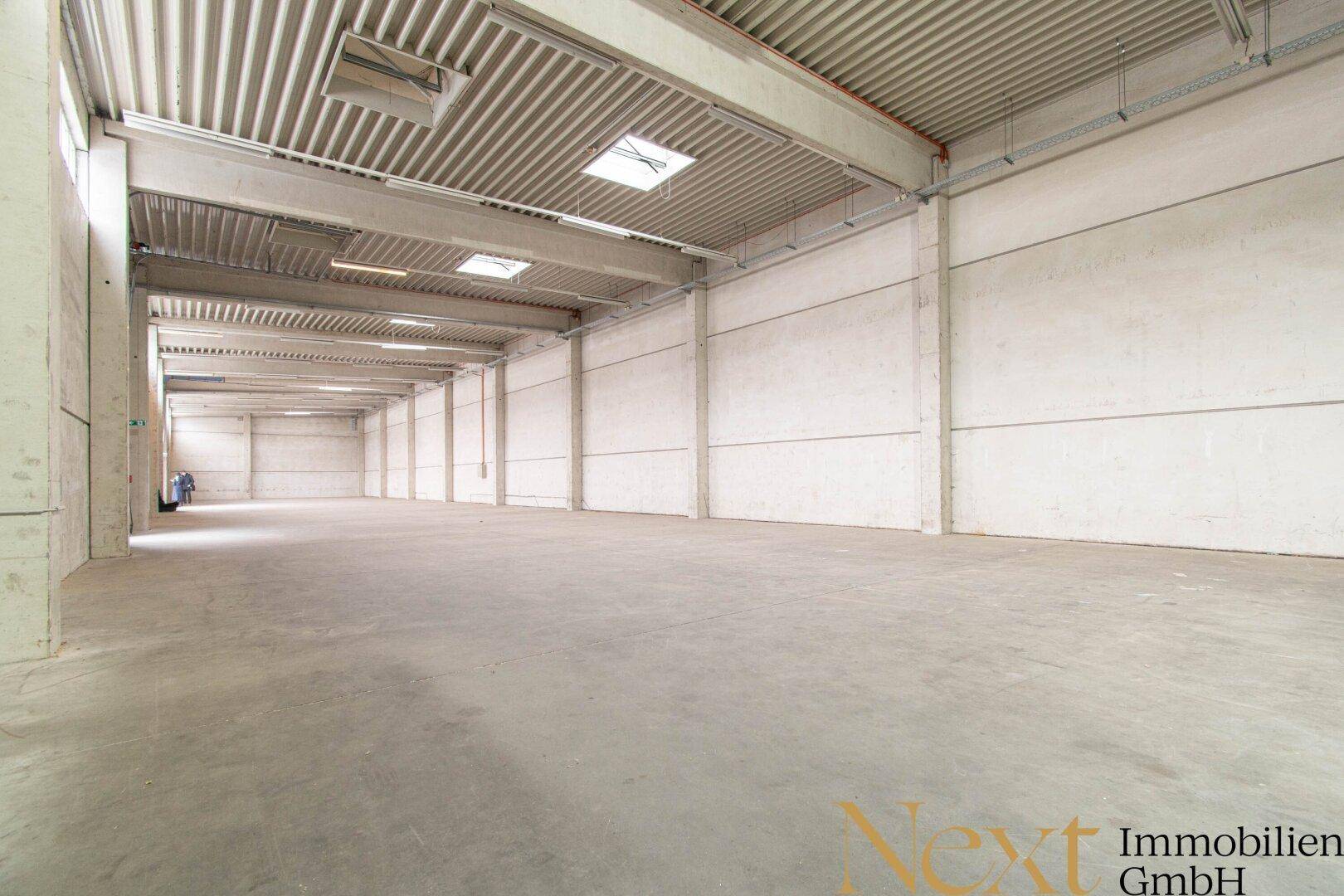 Lange Ansicht einer leeren Lagerhalle mit Betonboden, hohen Decken und Oberlichtern für Tageslicht.
