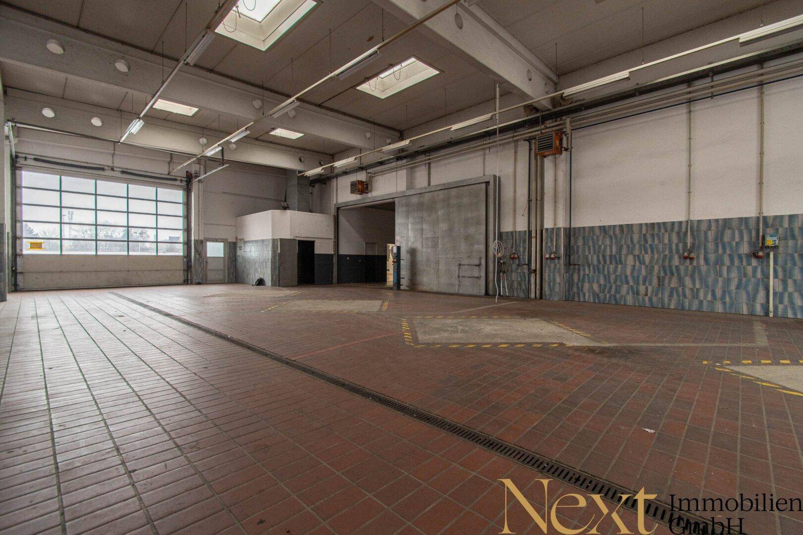 Werkstatt oder Garage mit roten Fliesen, hohen Decken und einem großen Glasrolltor für viel Licht.