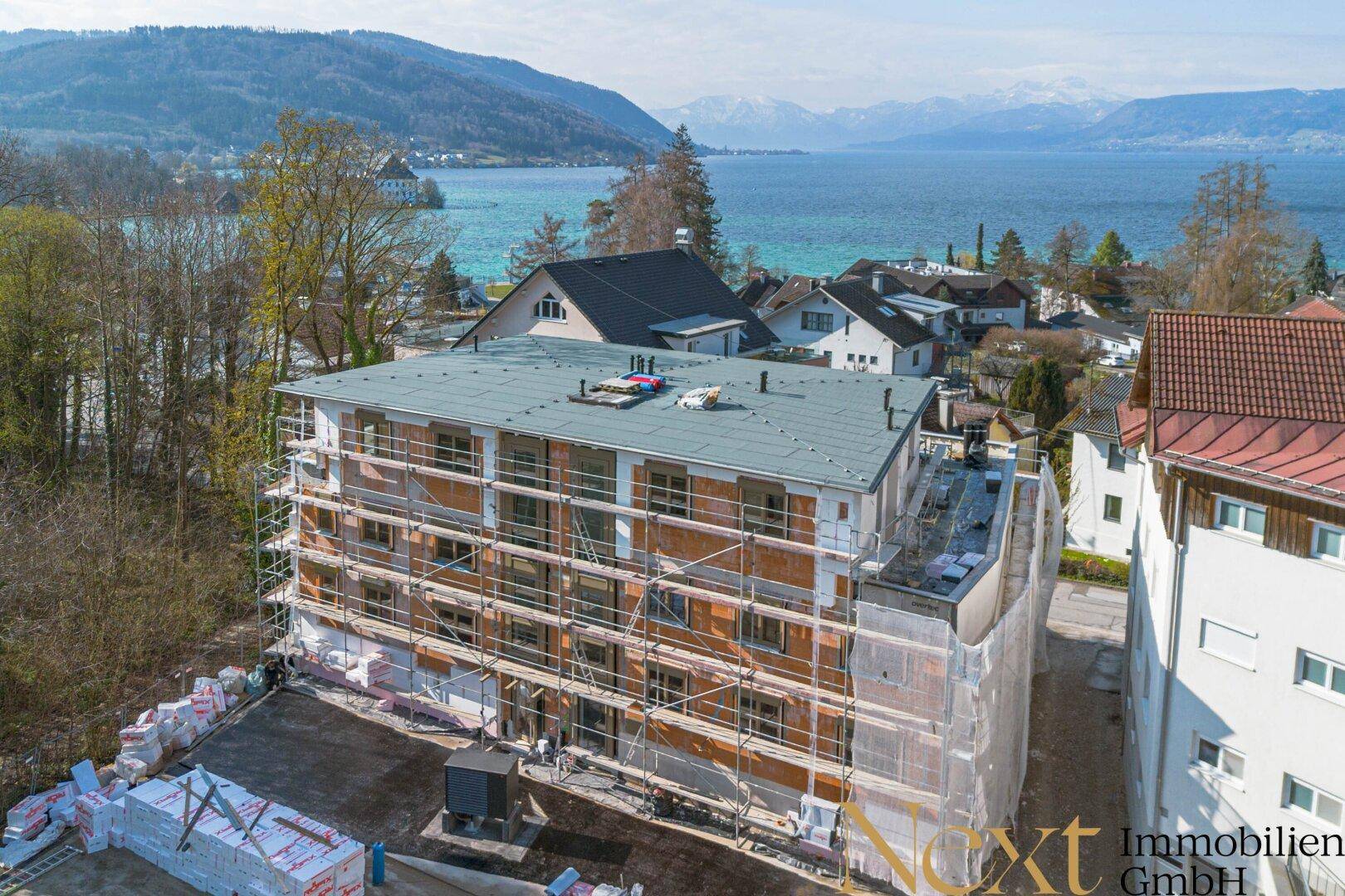 Luftaufnahme eines im Bau befindlichen Gebäudes mit Gerüst, direkt am See gelegen und mit Bergblick.