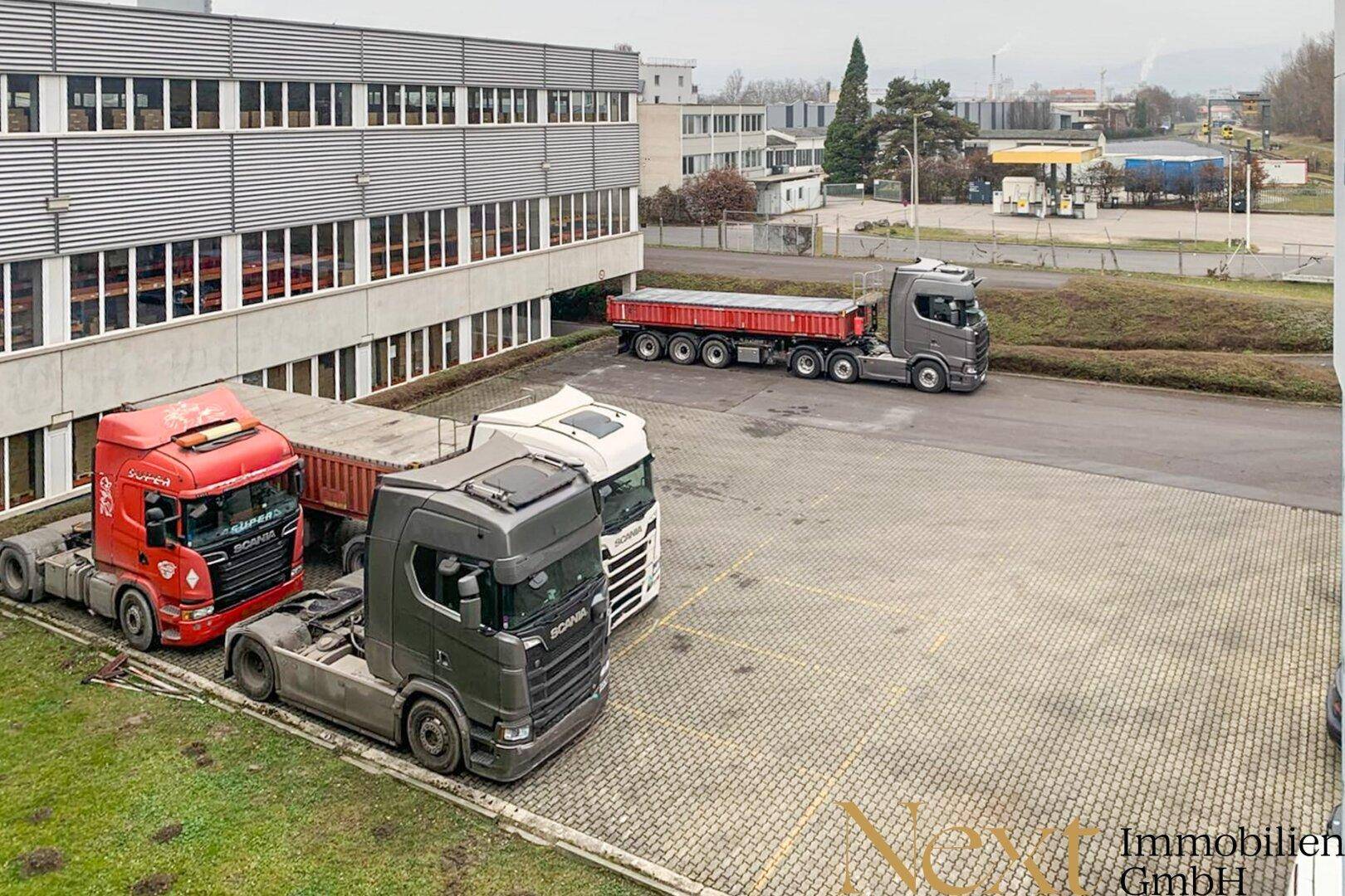 Außenansicht eines modernen Industriegebäudes mit großem gepflasterten Parkplatz und abgestellten Lastwagen.