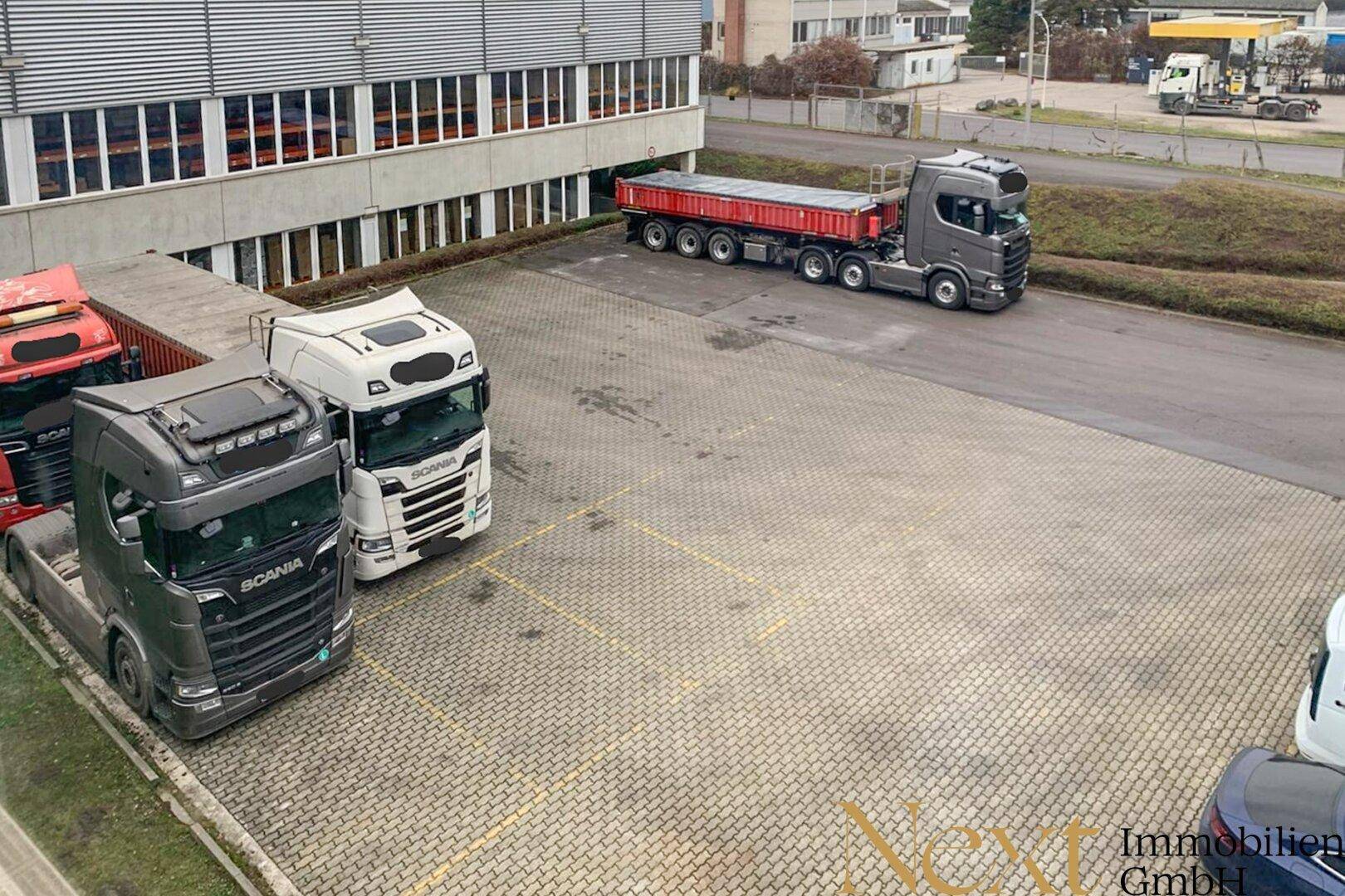 Industriegebäude mit moderner Fassade und großem Vorplatz, genutzt als Parkfläche für Lastwagen und Fahrzeuge.