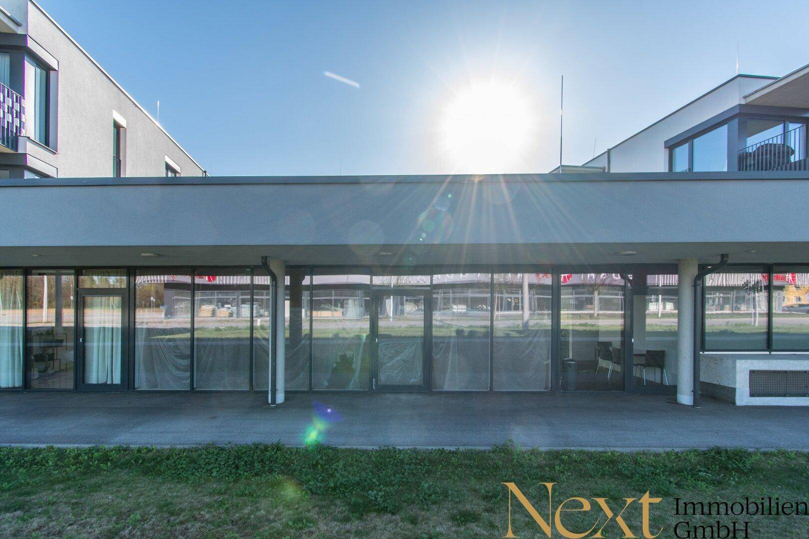 Sonnenbeschienene Gebäudefassade mit großen Fensterfronten im Erdgeschoss, die potenzielle Geschäftsräume zeigen.