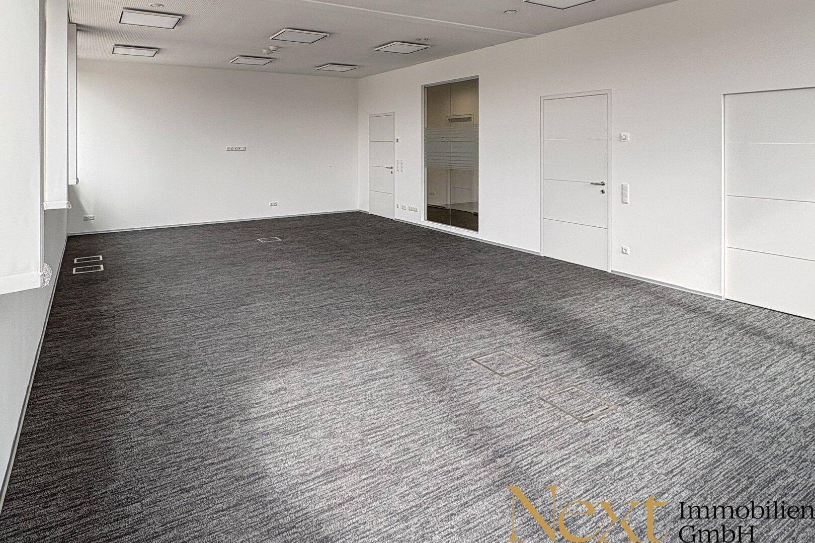 Leeres Büro mit grauem Teppichboden, weißen Wänden und mehreren Türen, bereit zur individuellen Gestaltung.