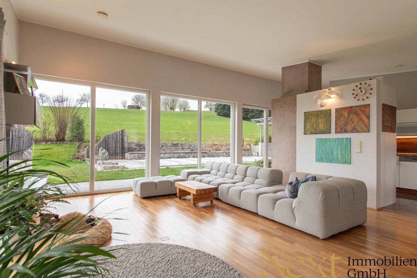 Geräumiges Wohnzimmer mit beigem Sofa, Holzboden und Panoramafenstern mit Blick auf die Natur.