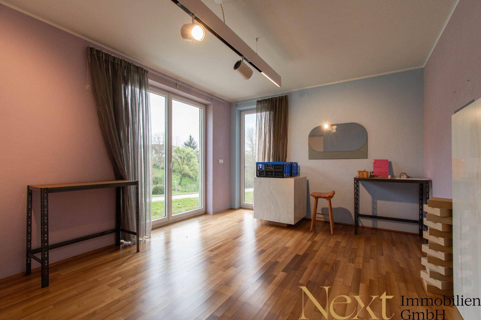 Helles Zimmer mit Holzboden, großen Fenstern und Blick ins Grüne, ideal als Wohn- oder Arbeitsbereich.