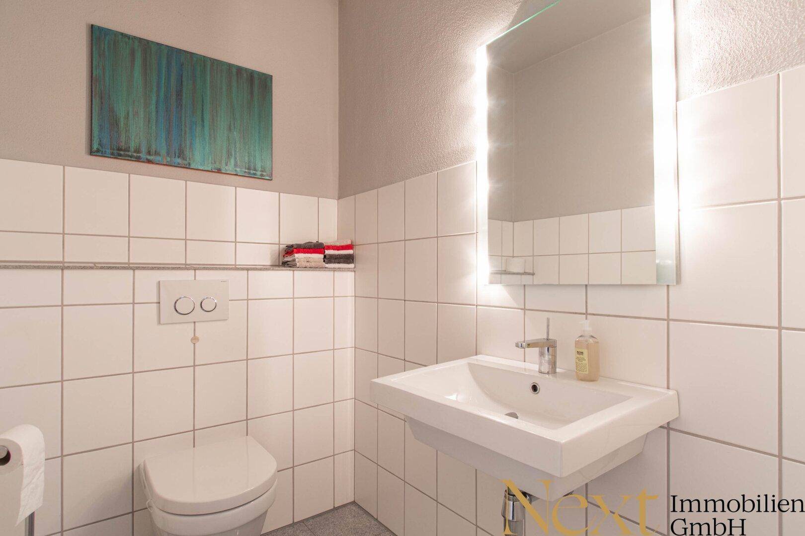 Modernes Gäste-WC mit weißen Fliesen, Wand-WC und beleuchtetem Spiegel über dem Waschbecken.