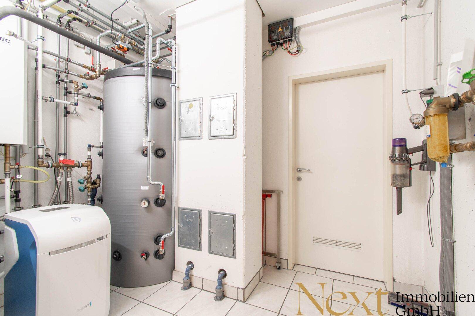 Heizungsraum mit großem Warmwasserspeicher, komplexer Rohrinstallation und weißen Bodenfliesen.