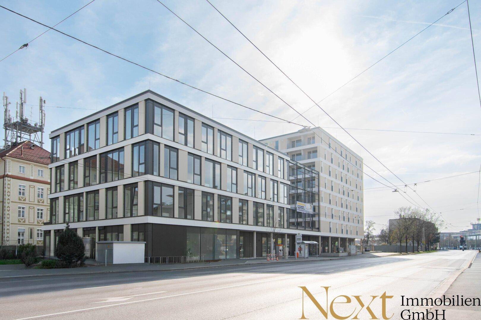 Weitläufige Ansicht eines modernen Geschäftsgebäudes mit angrenzendem Neubau in einer sonnigen Stadtlandschaft.