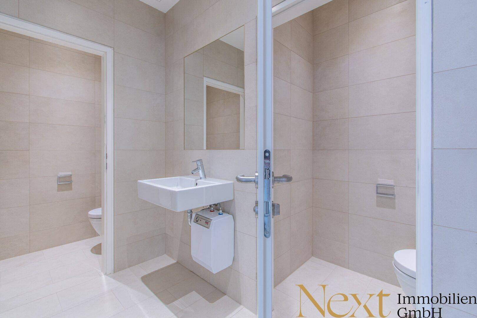 Helles Badezimmer mit beigefarbenen Fliesen, zwei Toiletten und einem modernen Waschbecken.