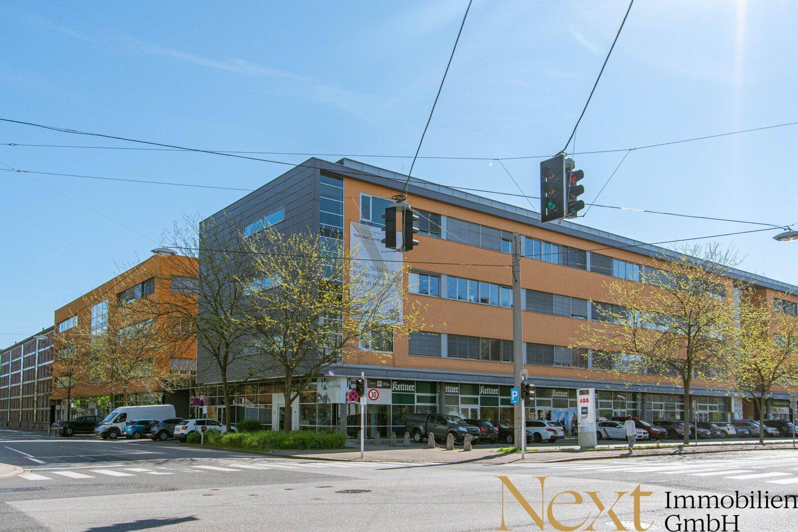 Modernes Bürogebäude mit orangefarbener und grauer Fassade an einer belebten Straße mit parkenden Autos und Bäumen.