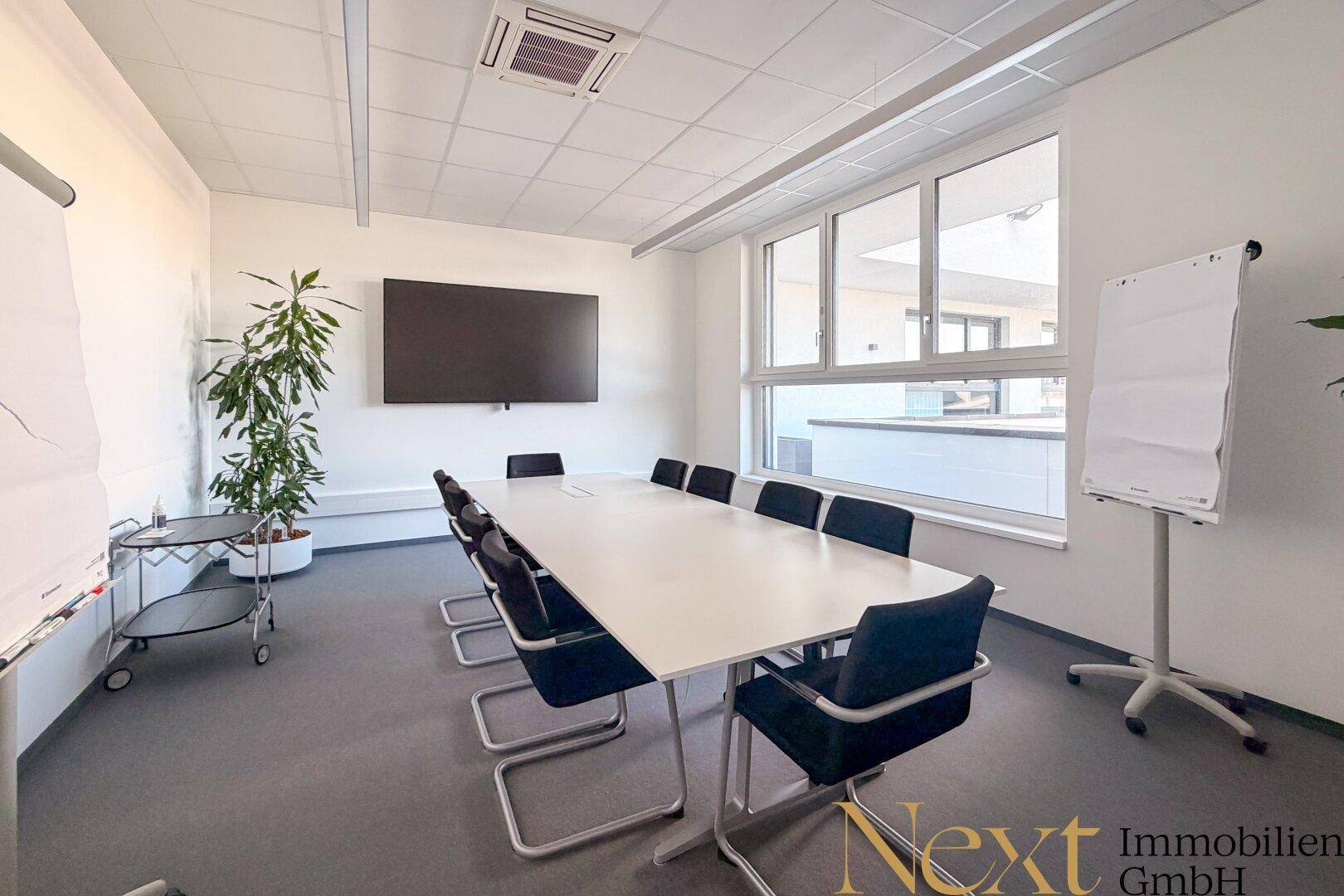 Moderner Konferenzraum mit großem Tisch, Stühlen, Wandfernseher und viel natürlichem Licht durch die Fenster.