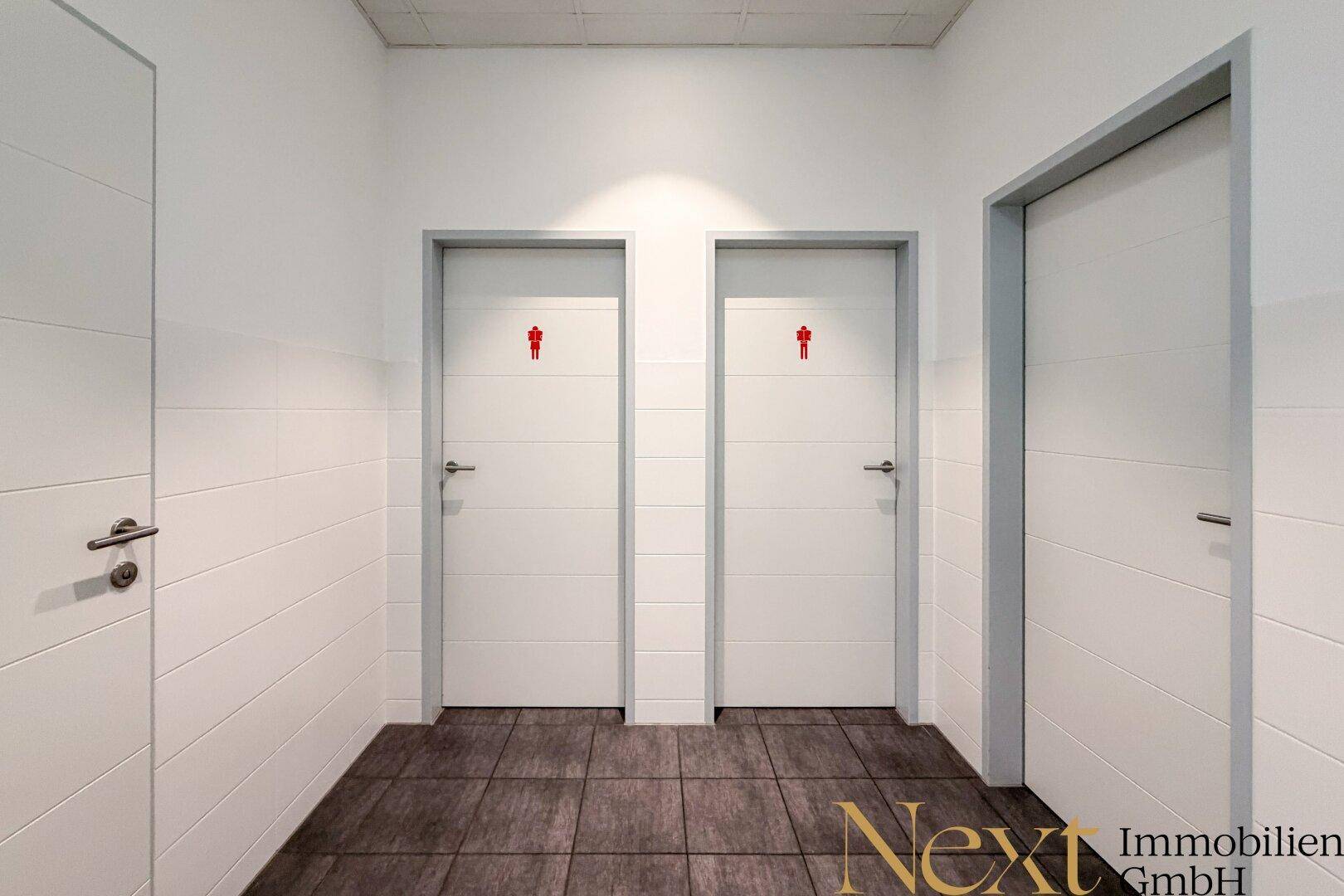 Heller Flur mit drei weißen Türen, zwei davon führen zu den Toiletten, mit dunklem Fliesenboden.