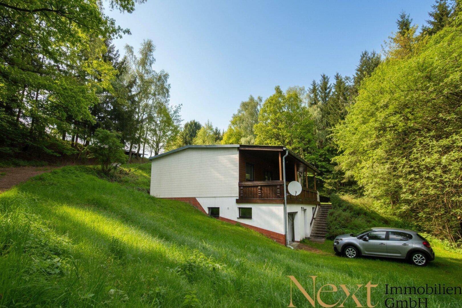 Freistehendes Haus in Hanglage, umgeben von viel Grün und hohen Bäumen, mit einer Zufahrt für Fahrzeuge.