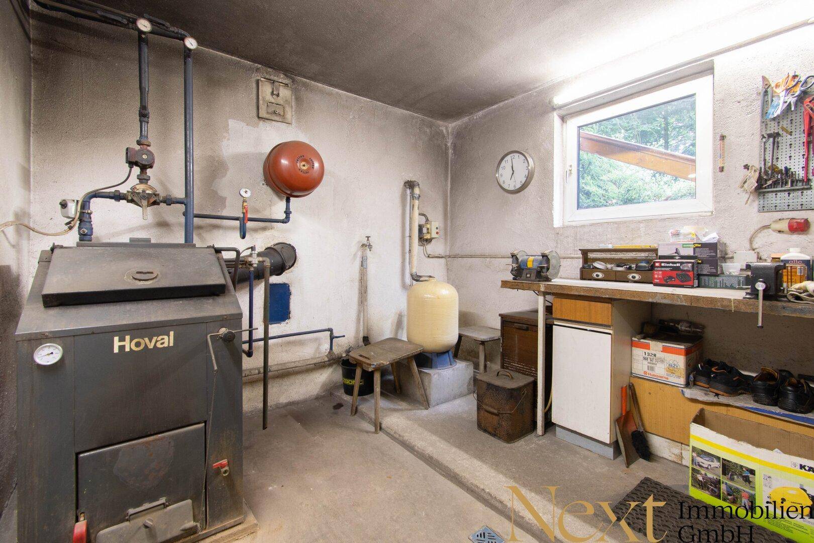 Funktionaler Heizungsraum im Keller mit Kessel, Rohren und einer Werkbank, gut beleuchtet durch ein Fenster.