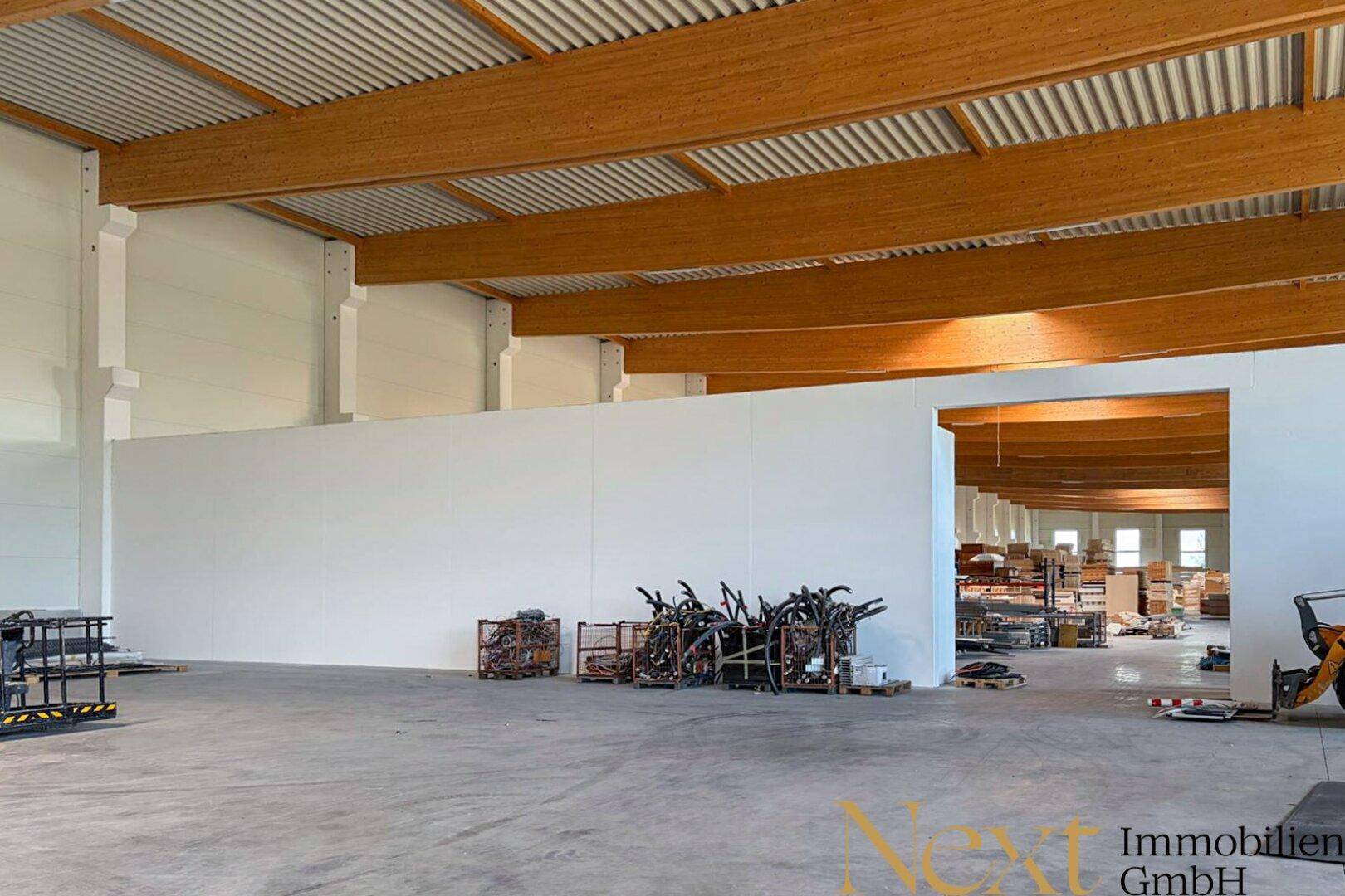 Geräumige Lagerhalle mit hohen Decken, sichtbaren Holzbalken und Betonboden, teilweise mit Trennwänden.