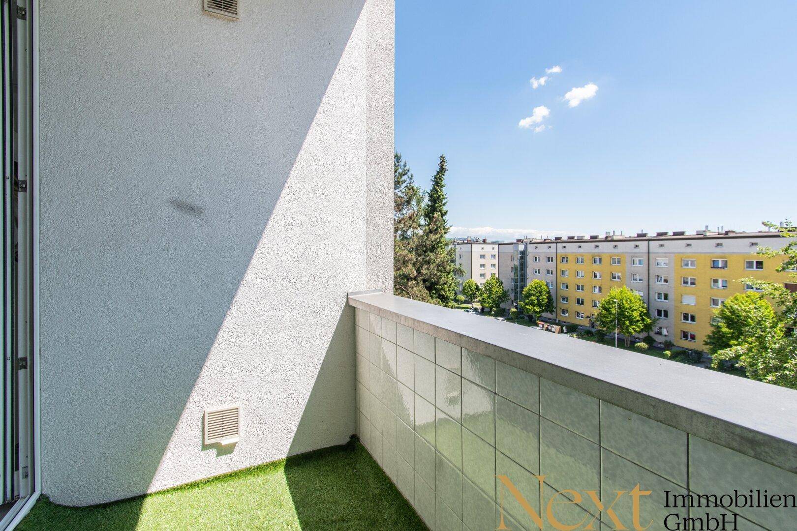 Kleiner Balkon mit Kunstrasen und Blick auf die umliegenden Wohngebäude und grüne Bäume.